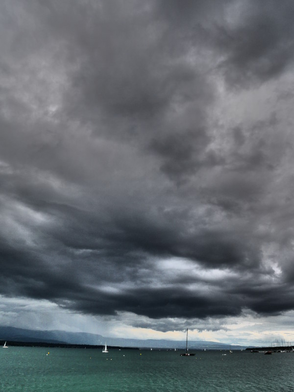 lac Léman, Genève, ciel orageux, nuages gris et lourds, lac émeraude avec quelques bateaux et une lumière à l'horizon, format portrait